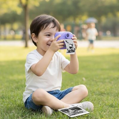 Petit garçon assis sur l’herbe dans un parc tenant un appareil photo instantané violet, souriant en regardant une photo imprimée posée devant lui.
